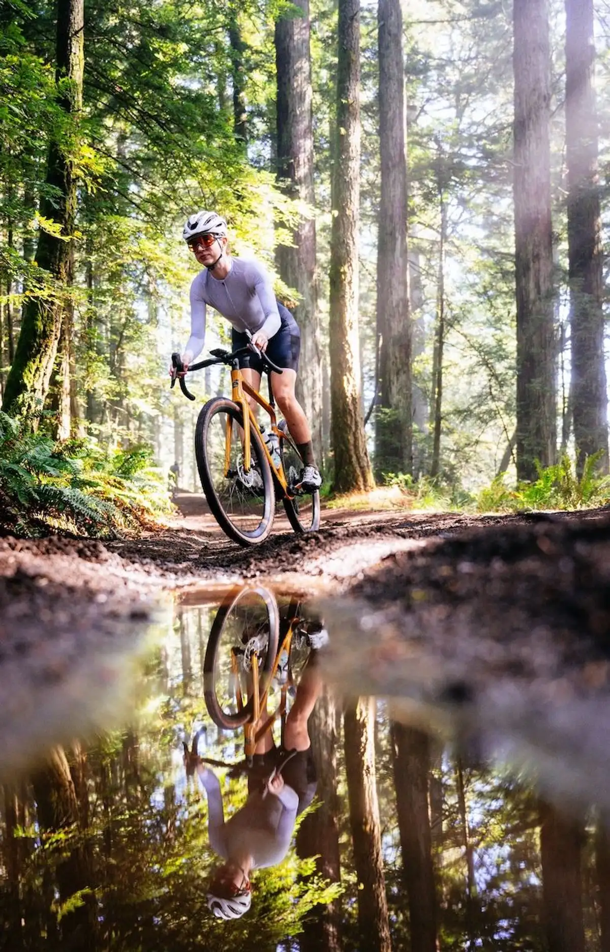 Frau fährt mit Gravelbike in sportlichen Klamotten durch Wald, vorne große Pfütze