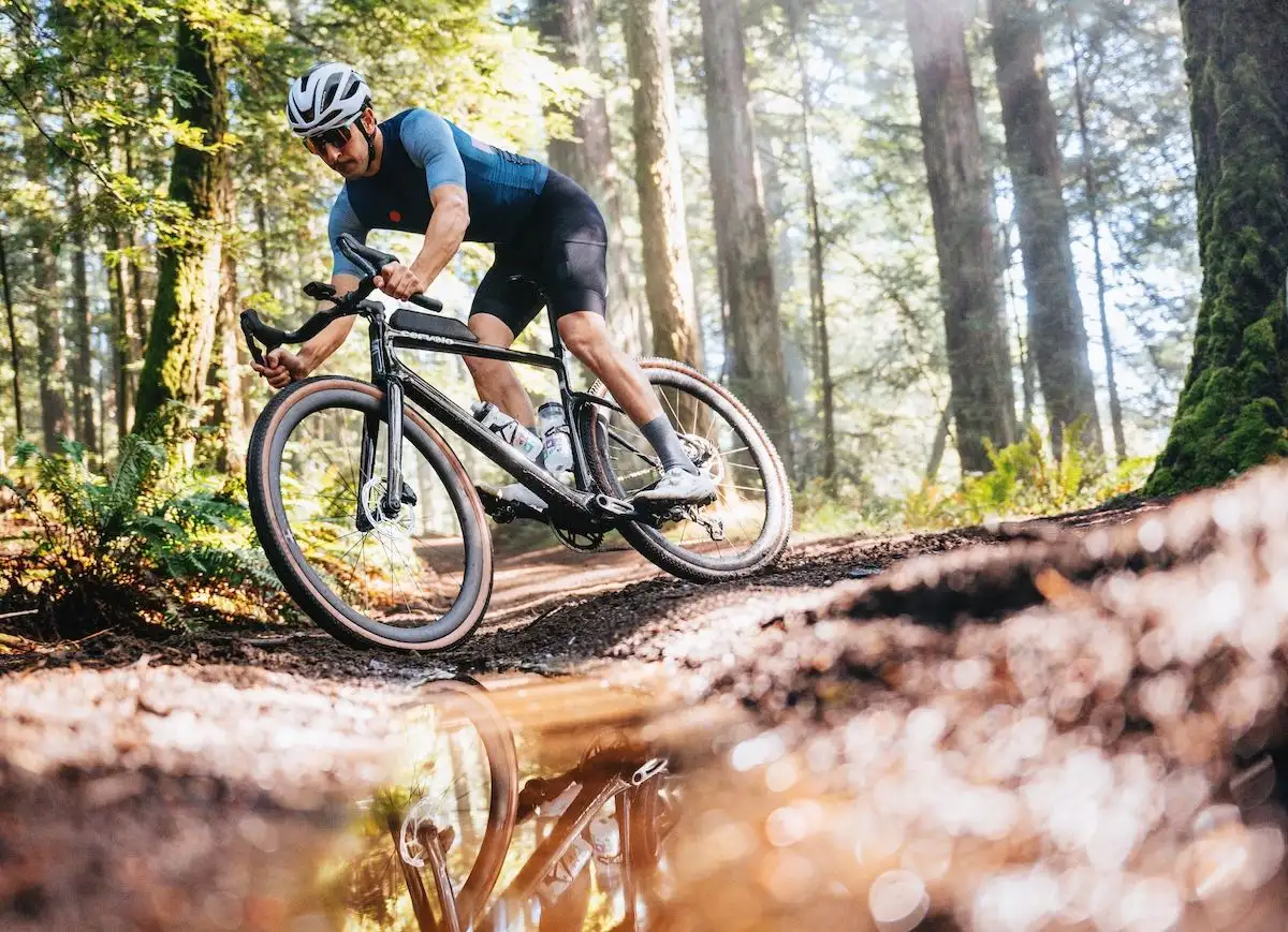 Man on a black gravel bike reflected in a puddle on a muddy forest path.