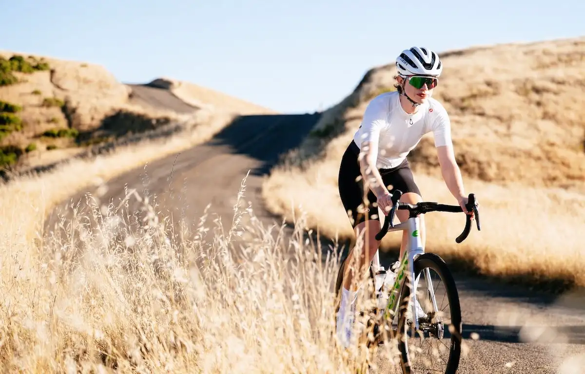 Woman in white and black cycling on a winding dirt road surrounded by dry golden grass.