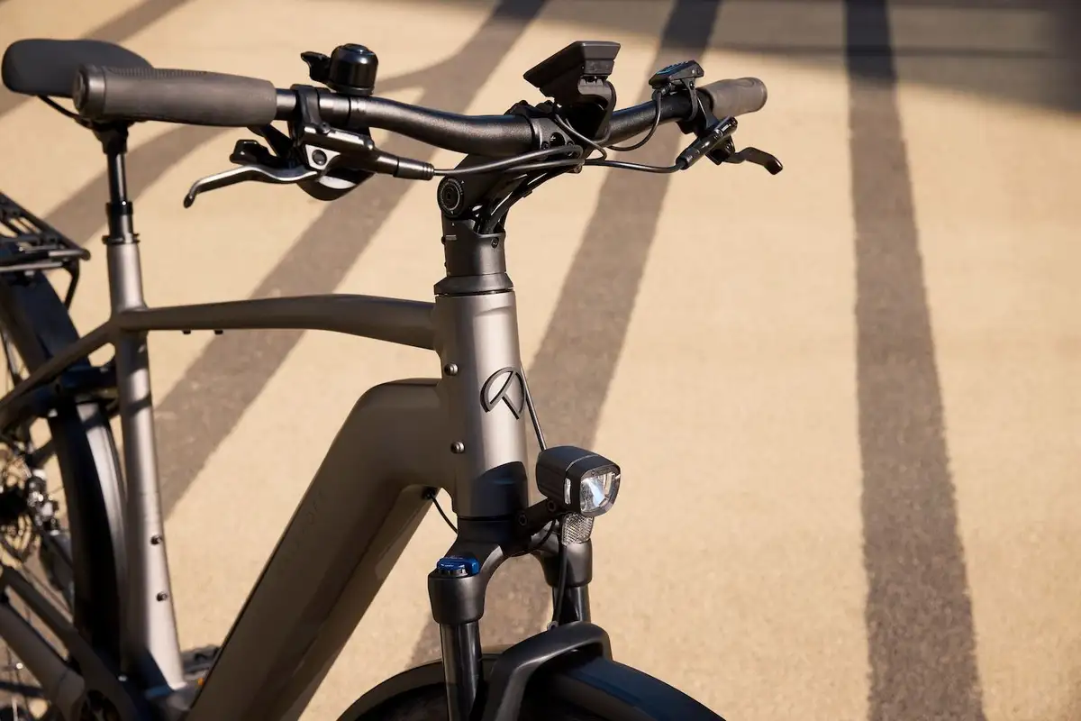 Close-up of a dark grey bicycle's handlebars, front light, and suspension in a city.