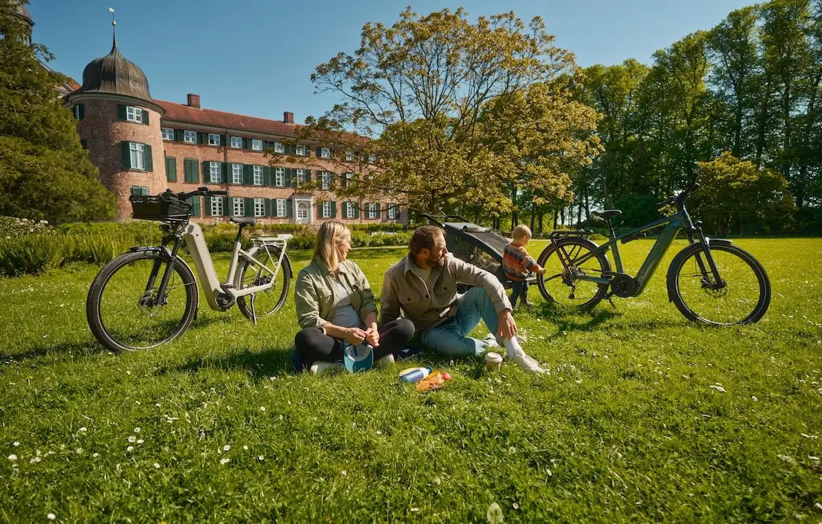 Two adults and a child enjoy a picnic on green grass with two bikes nearby.