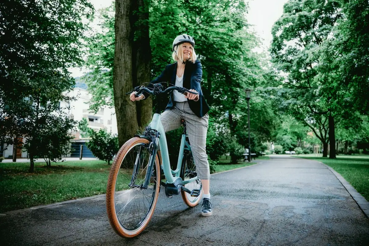 Frau mit Helm sitzt auf blauem E-Bike und stützt einen Fuß auf den Boden eines Radwegs umgeben von grünen Bäumen und schaut nach rechts