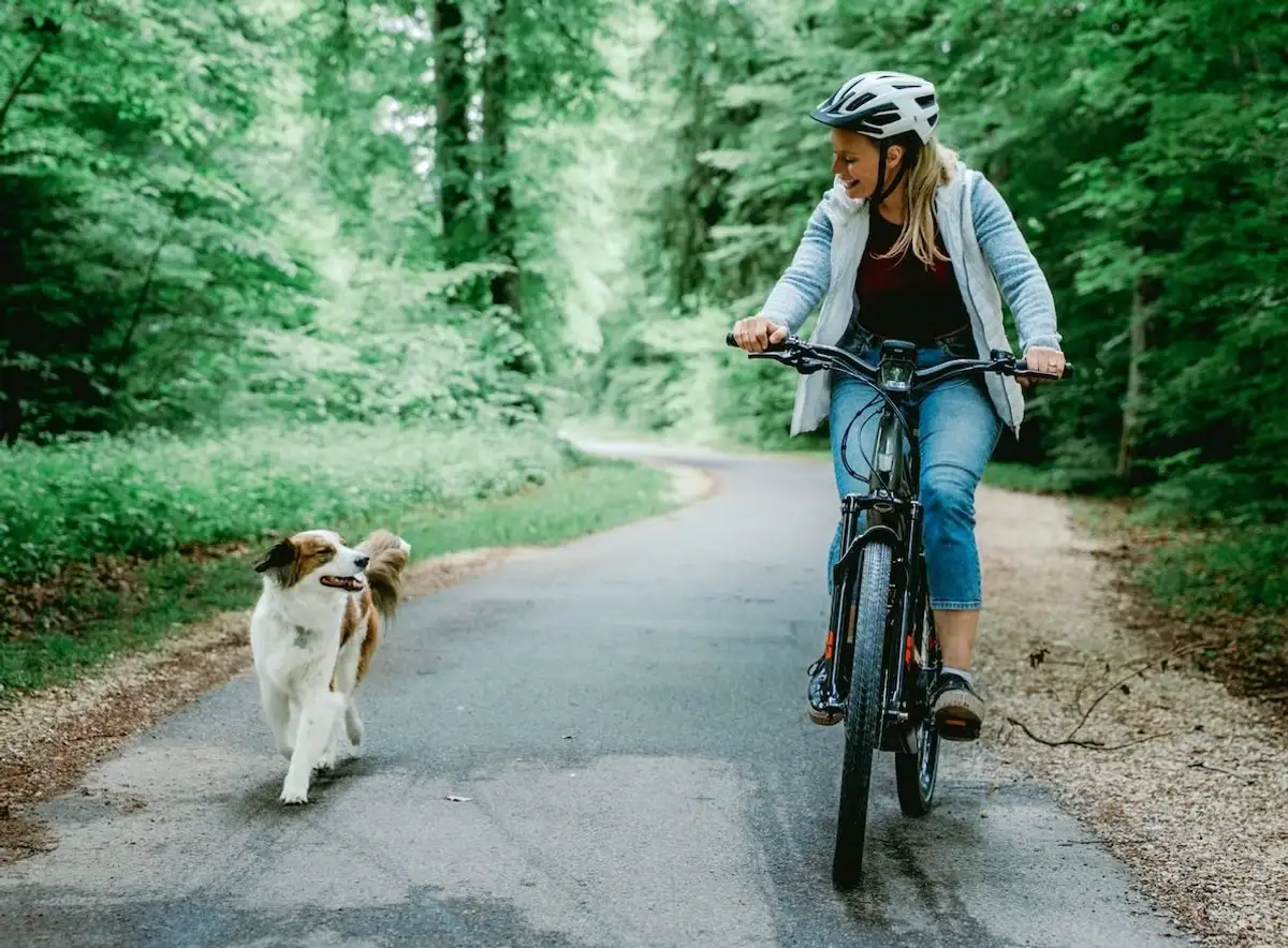 Woman in a helmet riding an e-bike, with a dog happily running alongside on a forest road.