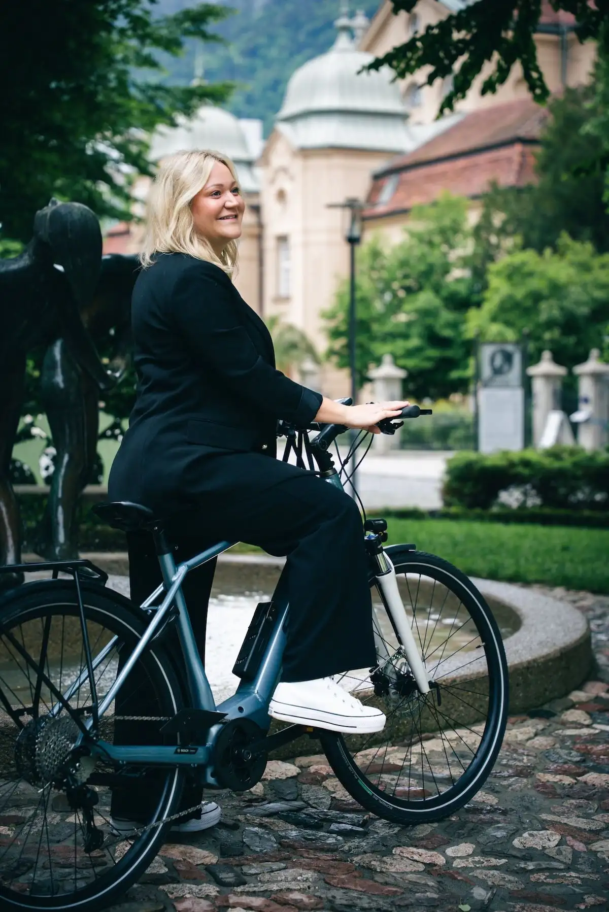 Blonde Frau in schwarzen Klamotten steigt lachend auf Dienstrad und steht auf Kopfsteinpflaster neben einem Brunnen, im Hintergrund alte Gebäude