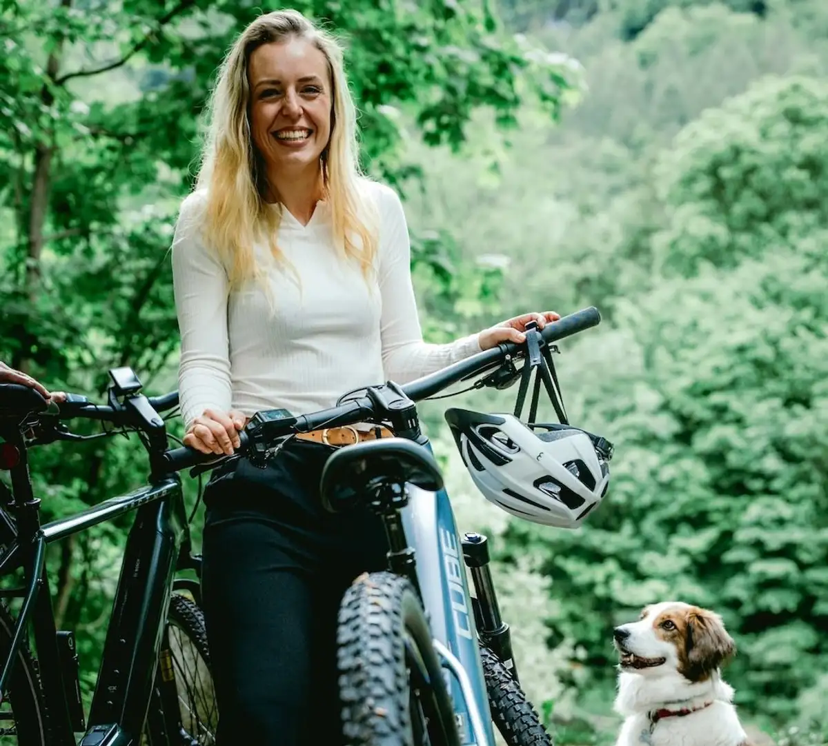 Junge blonde Frau steht neben Mountainbike und lacht in die Kamera, Hund sitzt daneben und lacht sie an