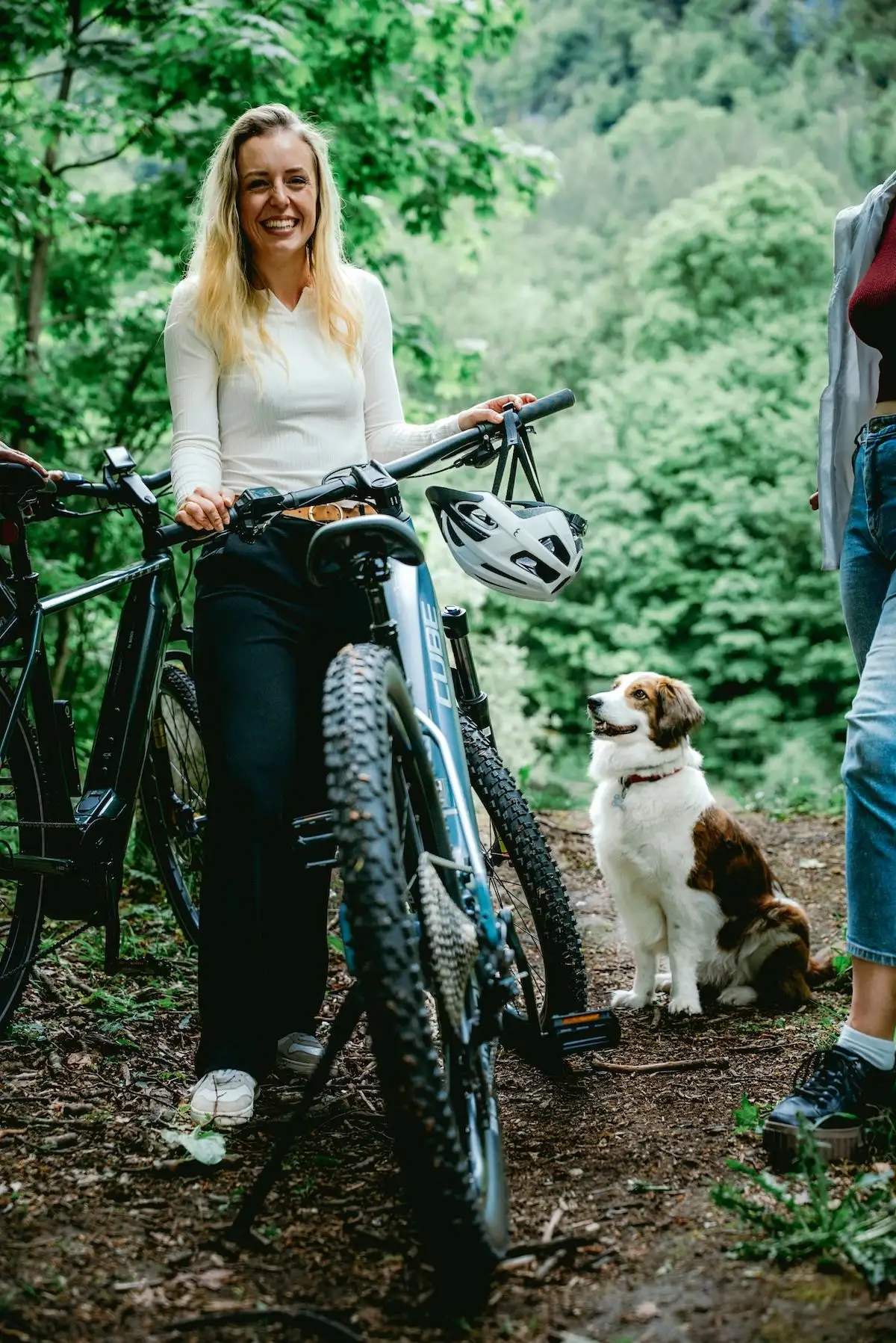 Frau steht mit Mountainbike lachend im Wald, Hund wartet daneben