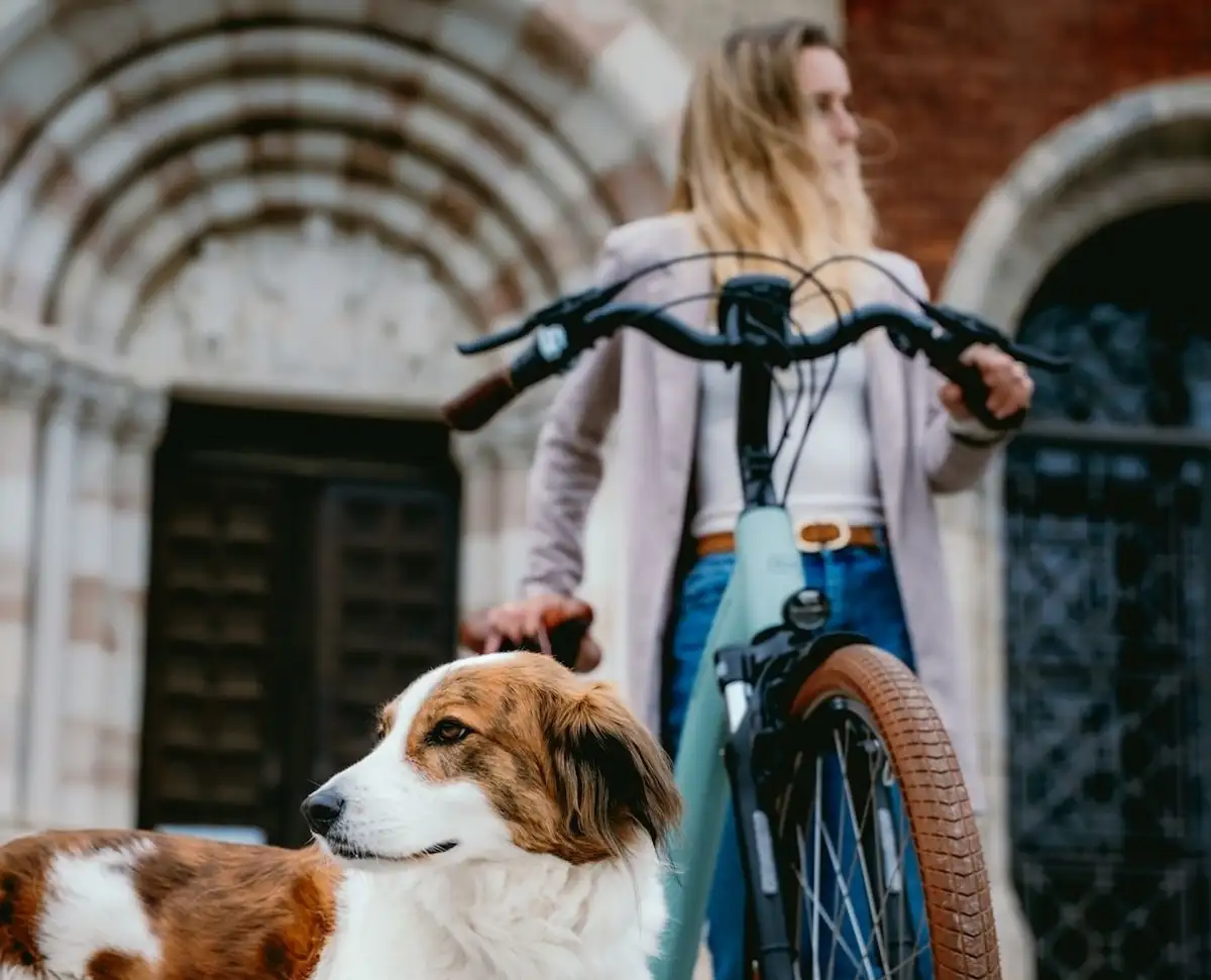 Frau steht mit Fahrrad im Hintergrund, vorne ist ein weiß-brauner Hund im Bild