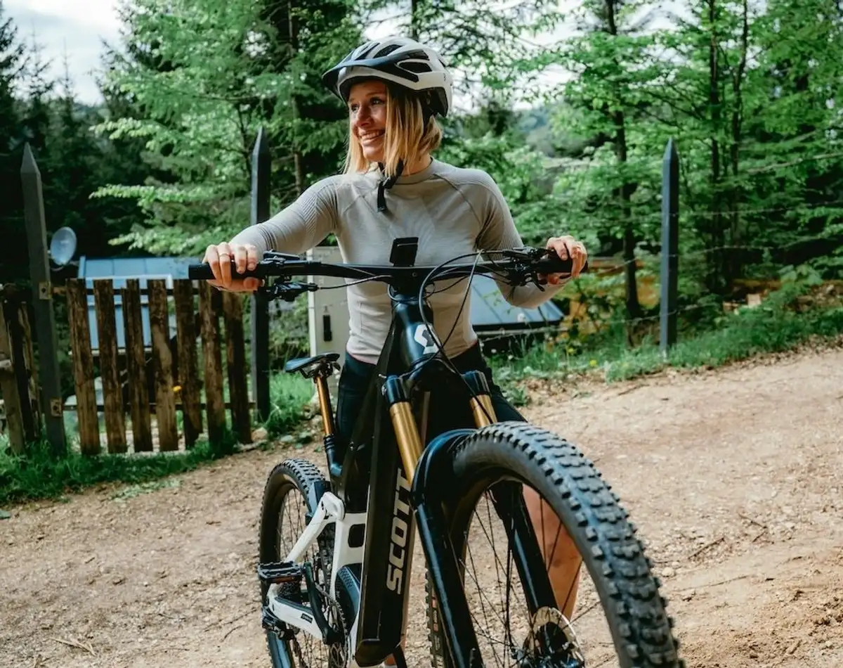 Frau mit Helm schiebt lachend Scott Mountainbike durch bergiges Waldgebiet