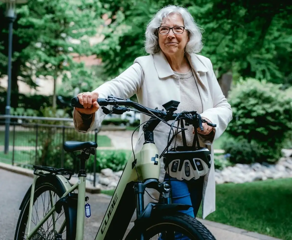 Ältere Frau schiebt e-Bike durch Park und schaut in die Kamera