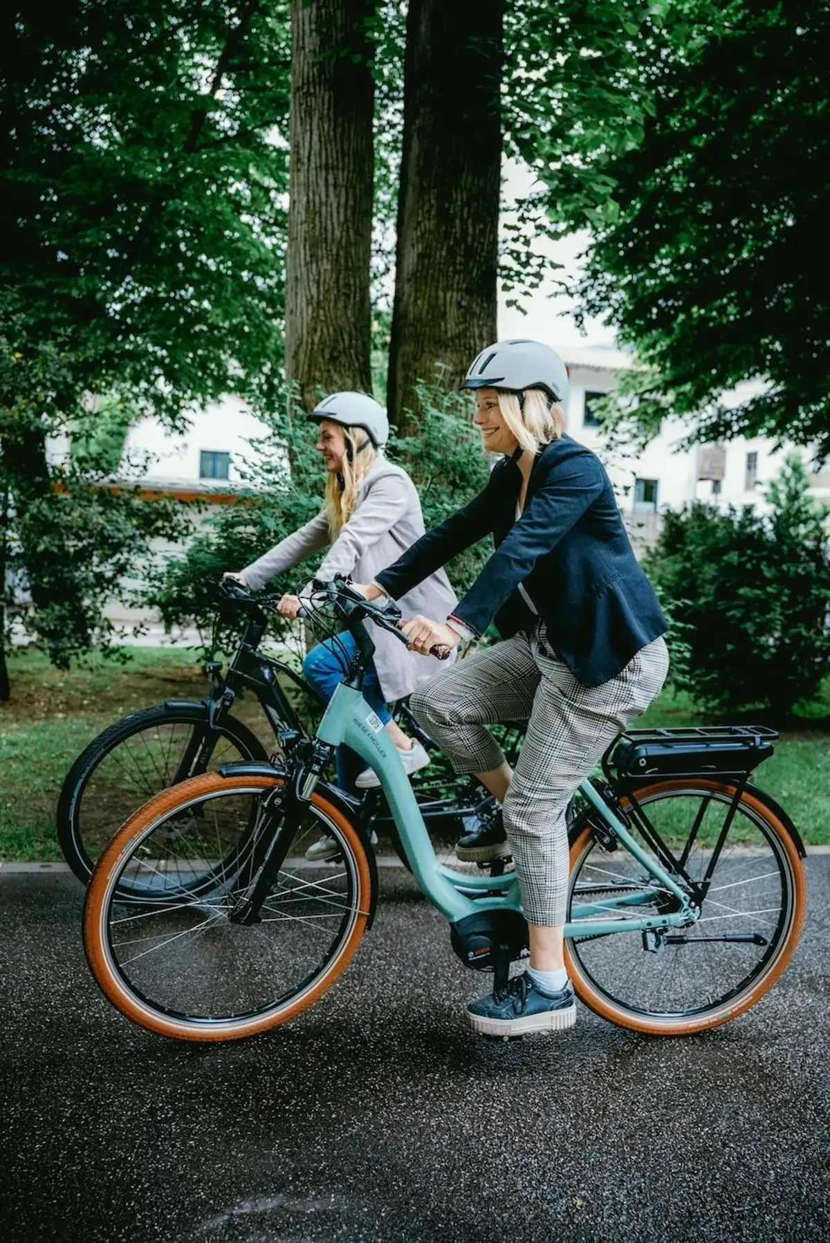 two women next to each other on her e-bikes smiling while riding a bike path in a city, in the background, there are green trees