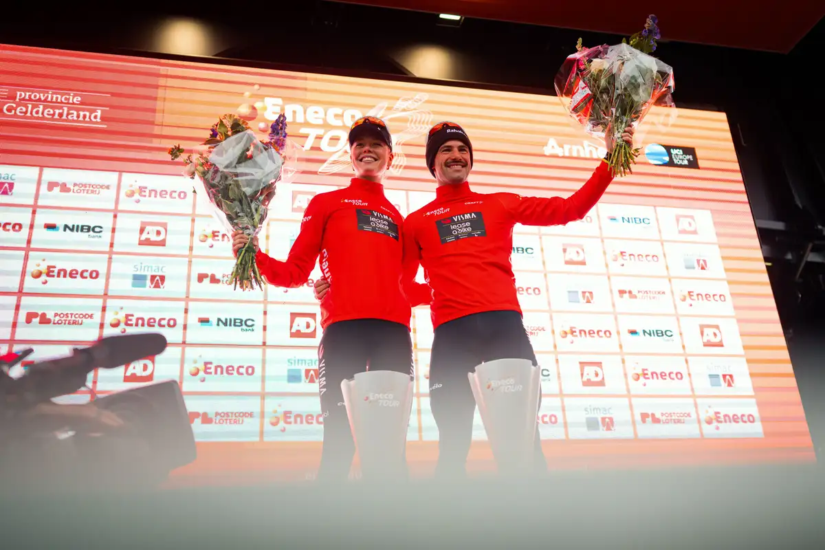 Two male cyclists in red jerseys stand on a podium, holding victory bouquets.