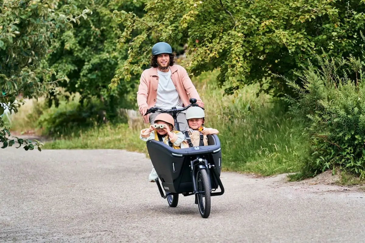 Mann mit Helm fährt zwei Kinder im schwarzen Lastenrad über Radweg entlang von grüner Natur