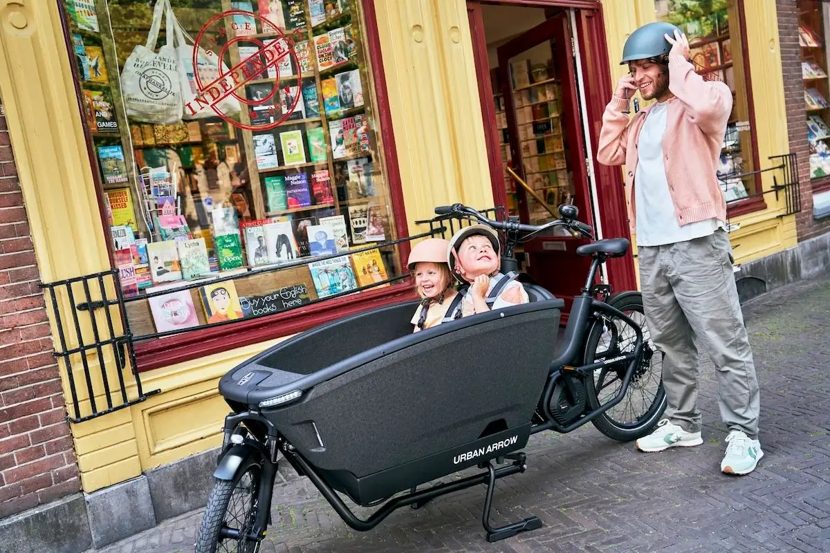Mann setzt gerade Helm und steht neben Lastenrad, Kindern sitzen vorne drin, ein Kind schaut Mann lachend über die Schulter an