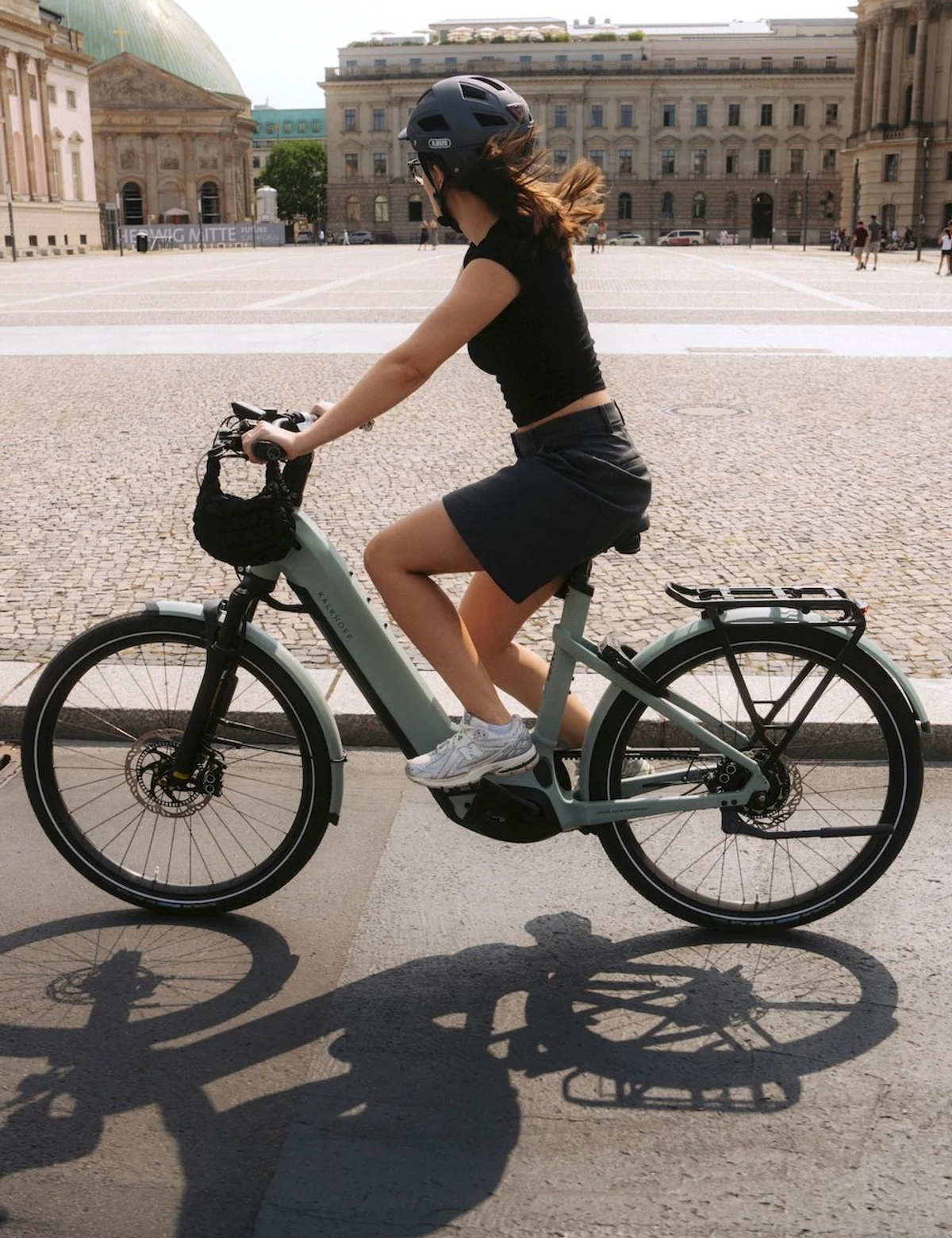 Frau in Sommerkleidung und mit Helm fährt mit E-Bike durch Stadt, kleine Tasche hängt am Lenker