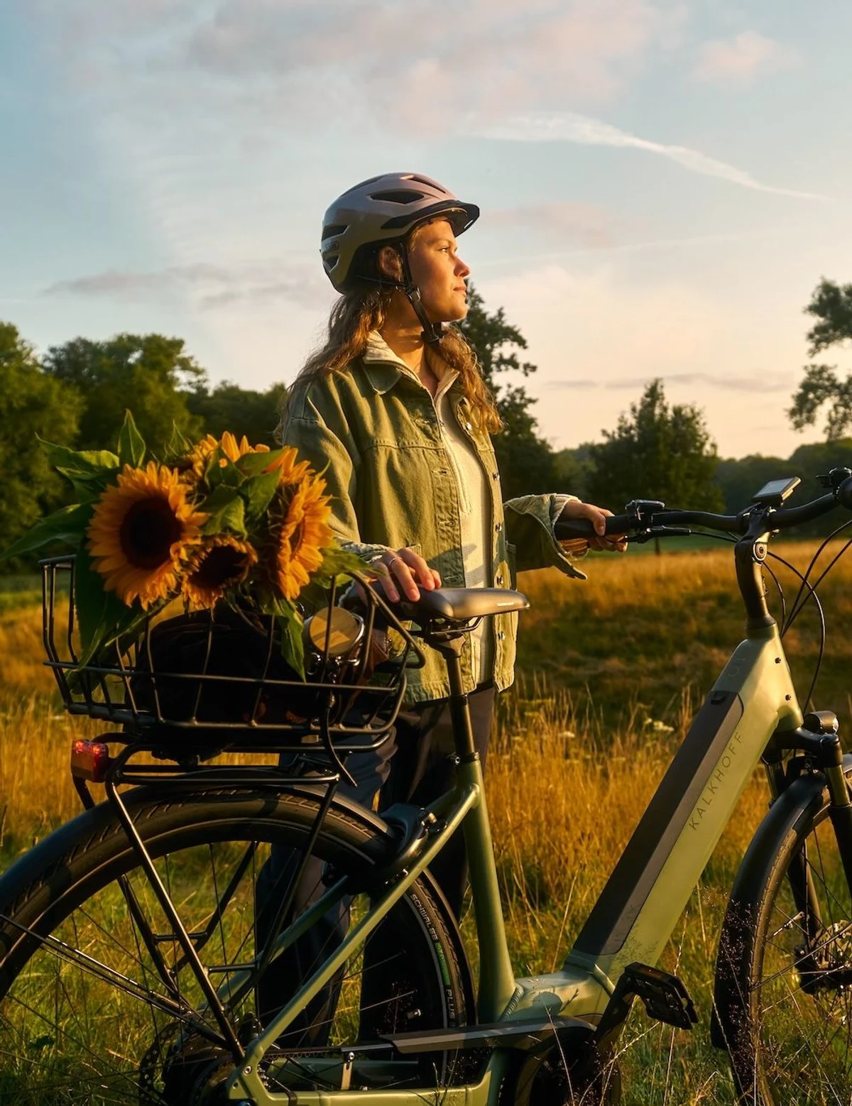 Frau mit Helm steht mit E-Bike und Sonnenblumen im Korb nahe Feld und schaut in den Sonnenuntergang