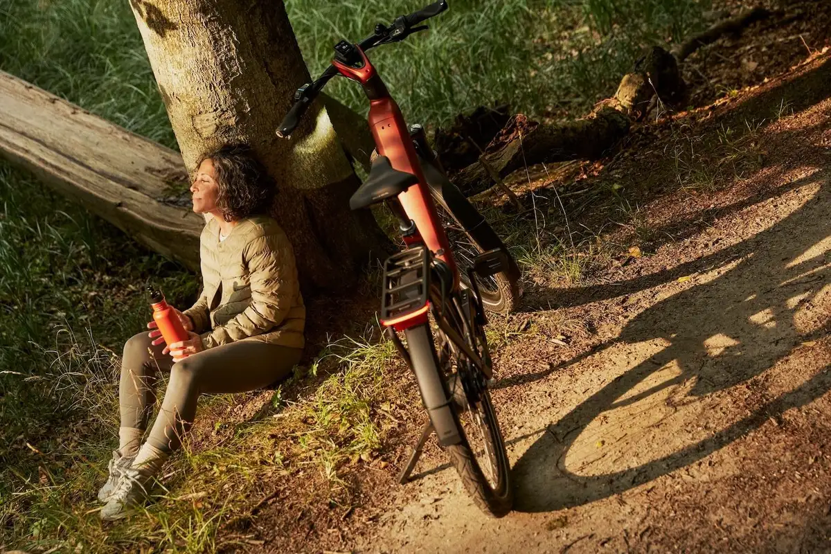 Frau sitzt an Baum und schaut Richtung Sonne, dahinter steht ein rotes Gazelle Fahrrad