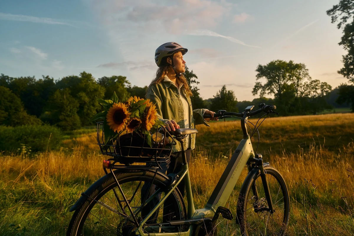 Frau mit Helm steht neben E-Bike in Sonnenuntergangslicht auf Feld und hat Sonnenblumen im Korb auf dem Fahrrad