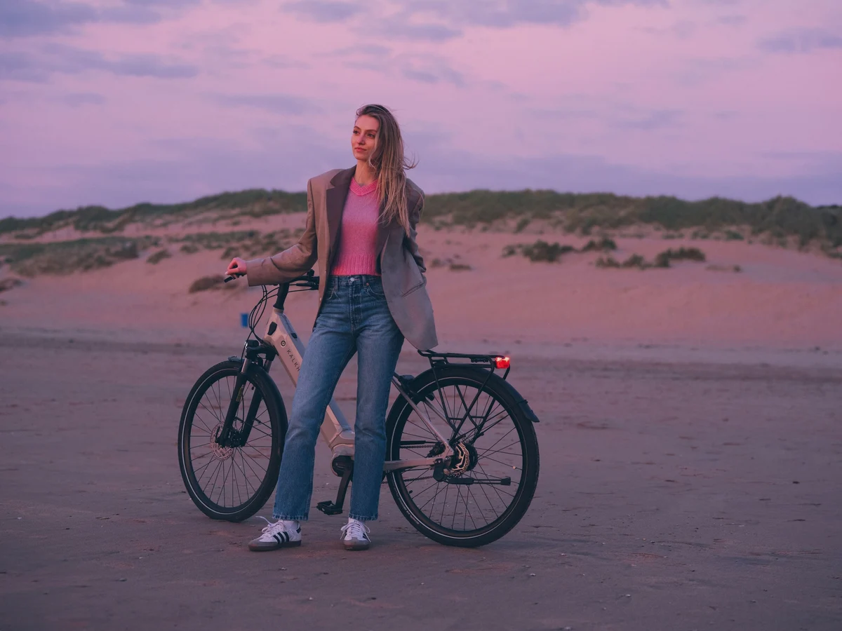 Woman stands with Kalkhoff e-bike on beach and looks at a pink-colored sky