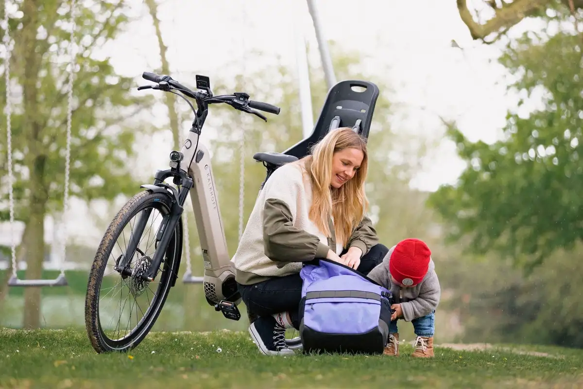 Frau mit Kind kramt in Rucksack Fahrrad im Hintergrund Kind sitzt neben Rucksack