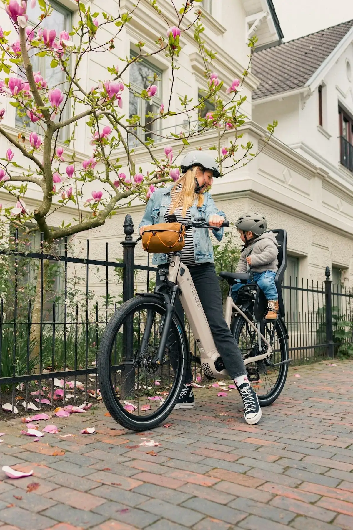 Young woman standing next to a fence with an e-bike, looking back at a toddler in a child seat