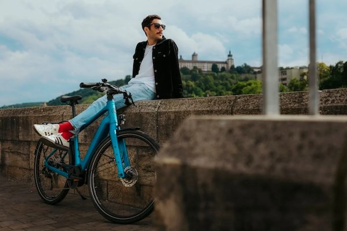 Mann mit Sonnenbrille und dunklem offenem Hemd und weißem Shirt und Jeans sitzt auf Mauer und schaut nach hinten, ein blaues Kalkhoff E-Bike steht vor ihm