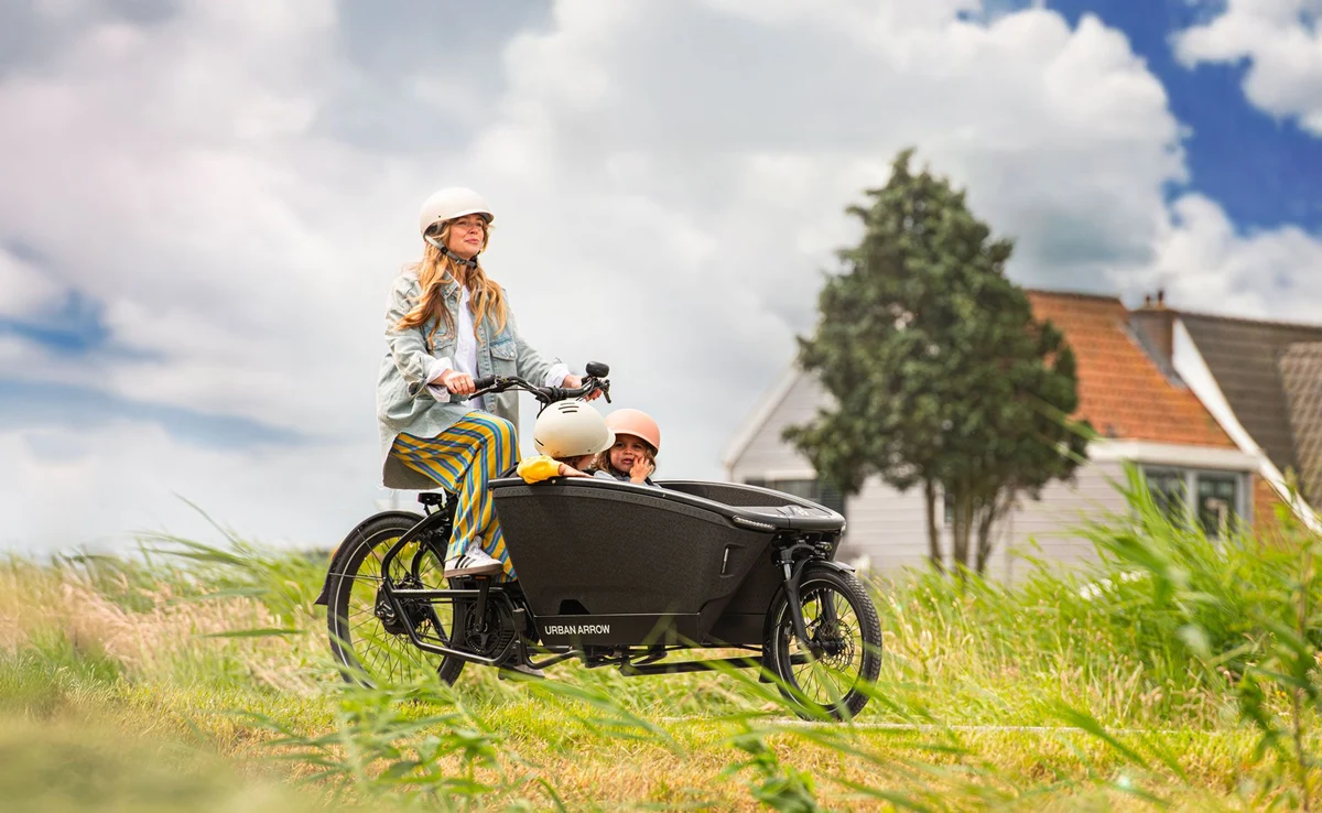 A woman rides a cargo bike with two children sitting in the front through a dune-like landscape; a house is visible in the background