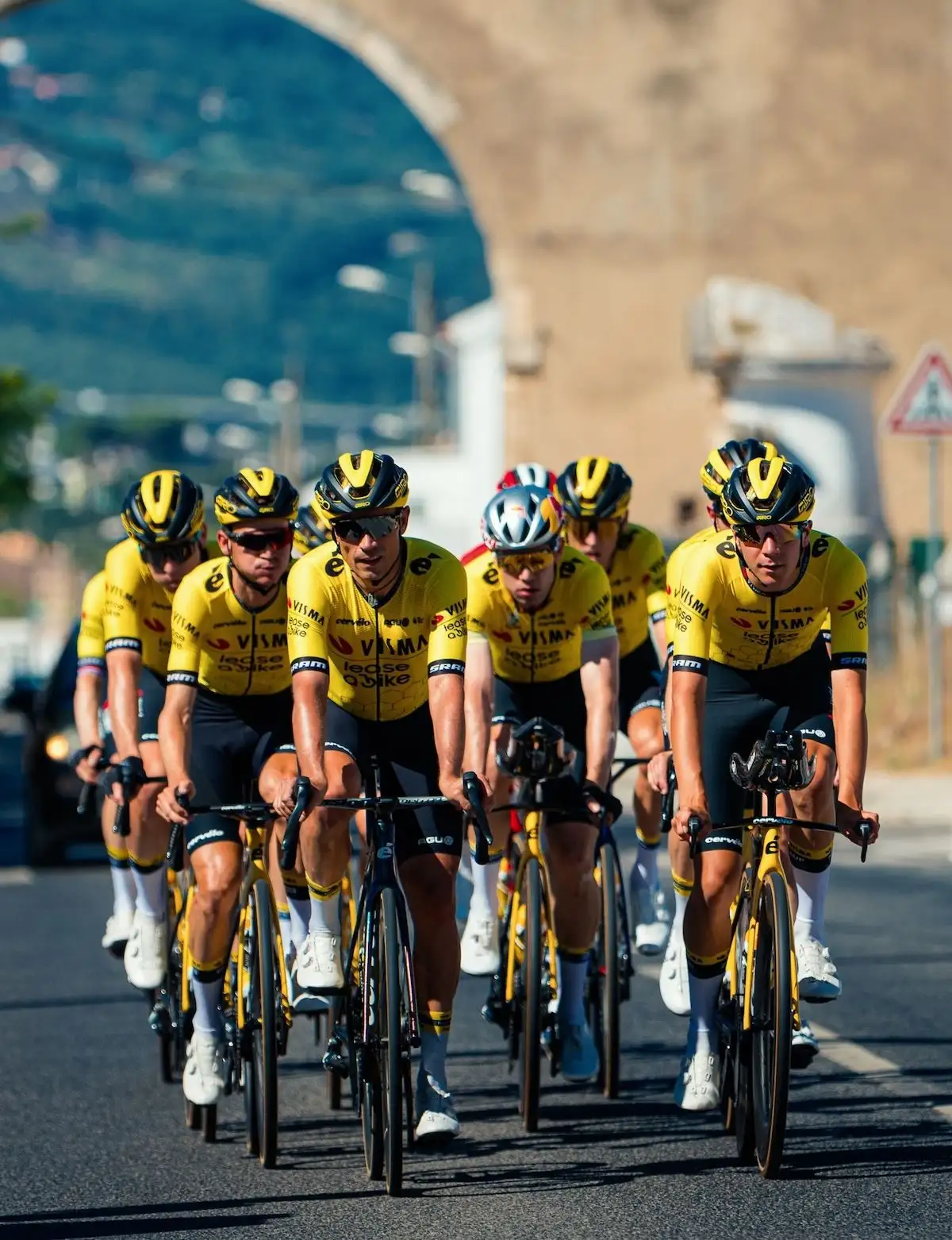 Team Visma Lease a Bike auf Rennrädern fahrend auf Straße vor Torbogen